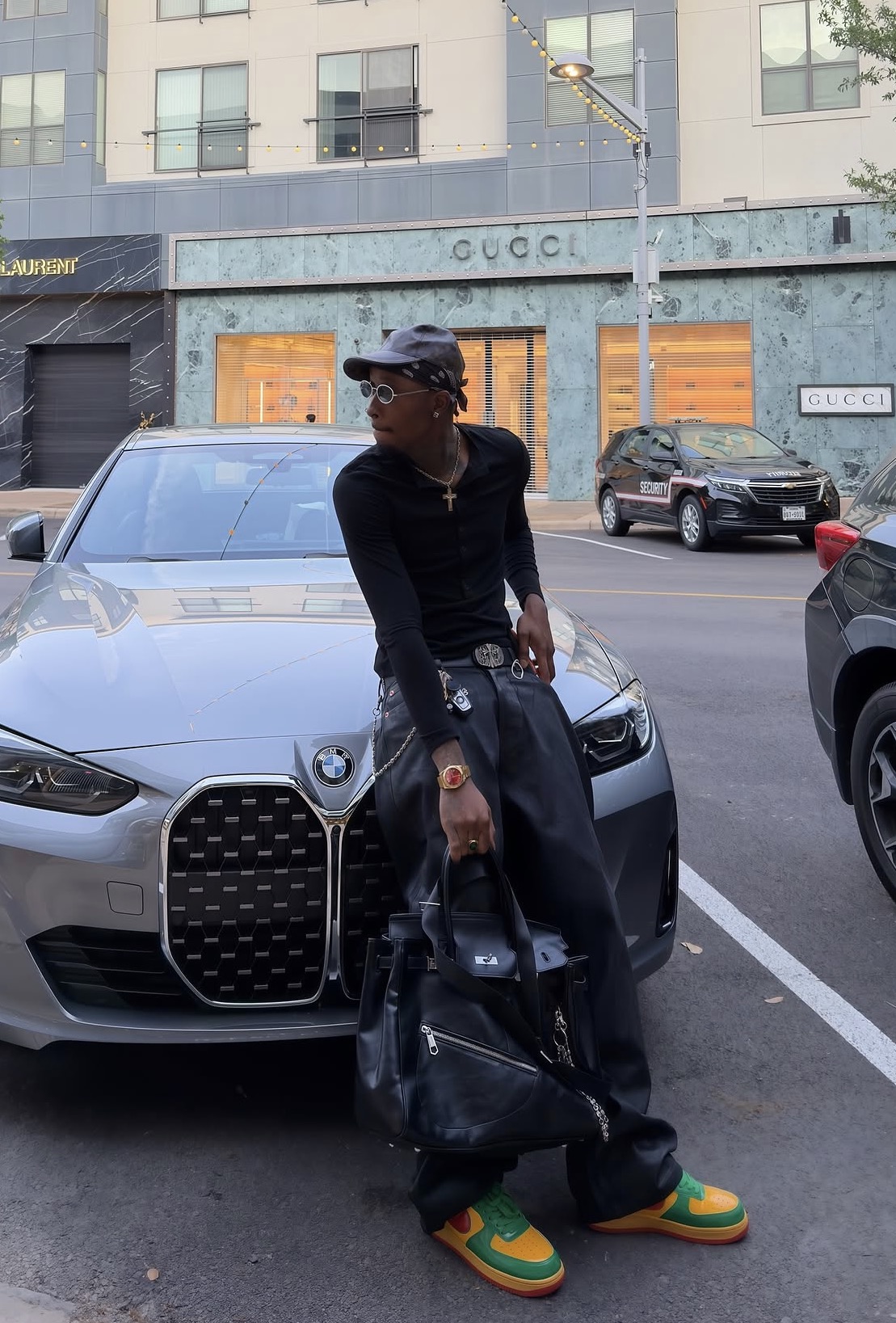 Claudio Conde in a black outfit with colorful sneakers and a leather bag beside a silver BMW